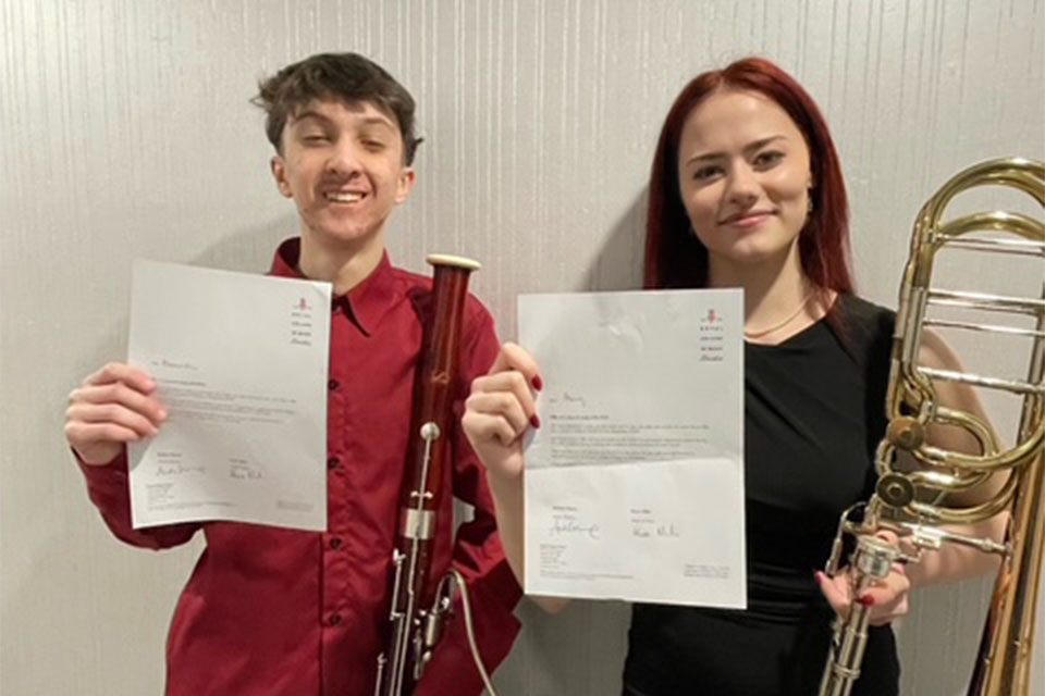 A young man and woman holding RCM acceptance letters smiling at the camera. The boy is holding a bassoon and the girl is holding a bass trombone.